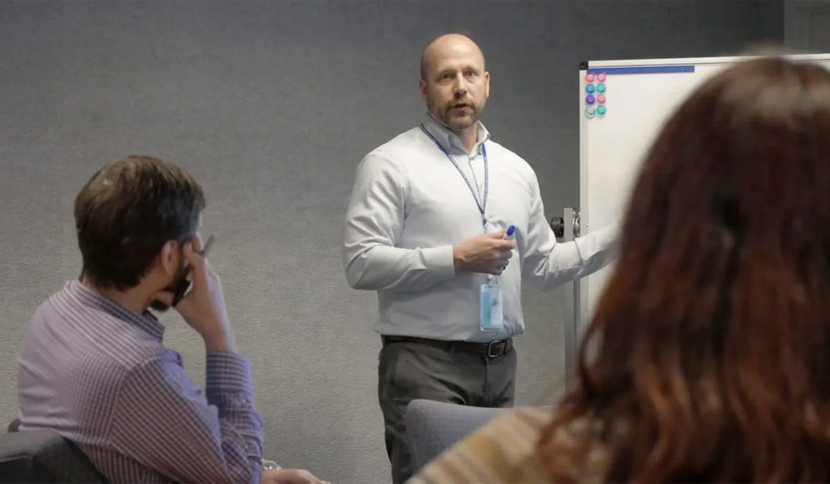 Systems Planning & Analysis employees meet and listen while the speaker explains a diagram on the whiteboard