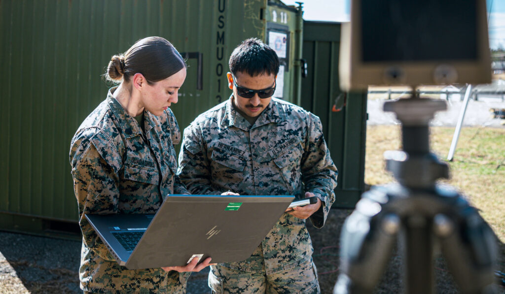 Two military members stand outside in the field reviewing a laptop screen