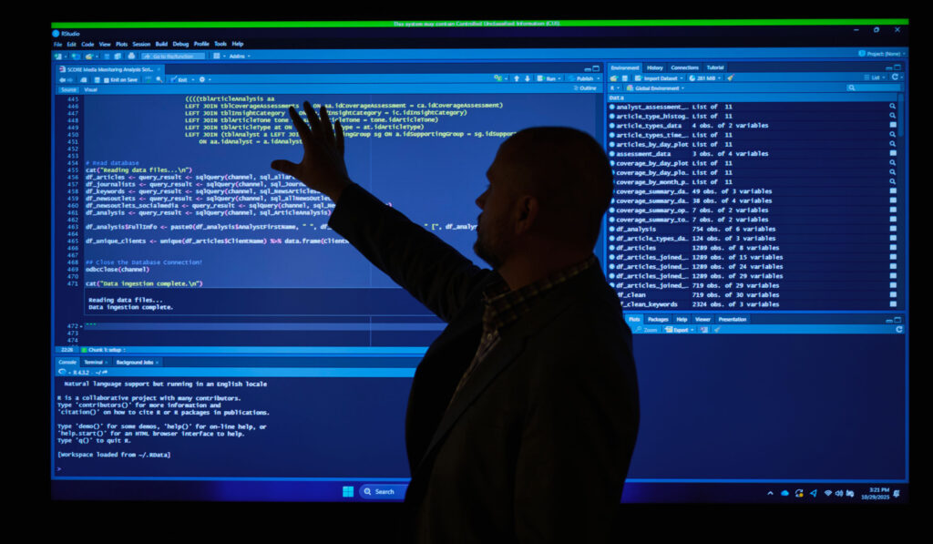 The silhouette of an SPA employee standing in front of a large screen showing RStudio Score script and code, depicting DevSecOps
