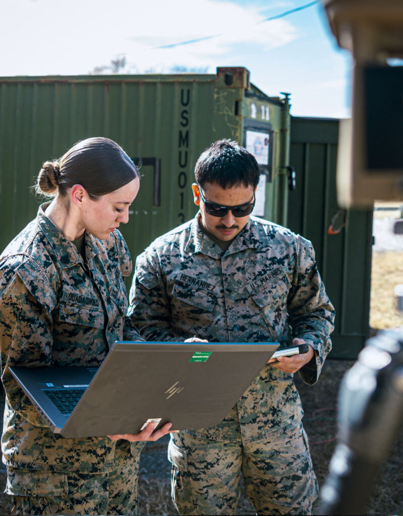 Two military members stand outside in the field reviewing a laptop screen