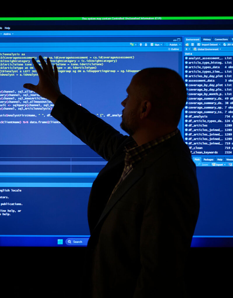 The silhouette of an SPA employee standing in front of a large screen showing RStudio Score script and code, depicting DevSecOps