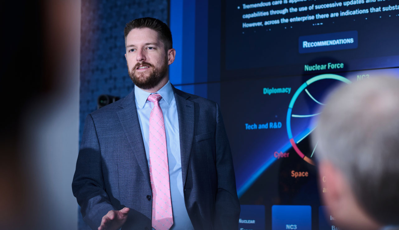 SPA employee leads a meeting with others on his team. The background screen shows SPA's Posture Commission Implementation System (PCIS) capabilities